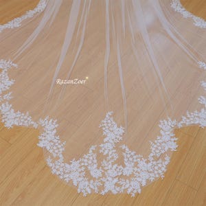 Puede incluir: Un velo de novia de tul blanco con un delicado borde de encaje floral. El velo está colocado sobre una superficie de madera.
