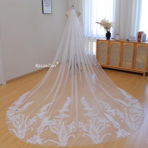 May include: A long, white bridal veil with a floral lace border. The veil is displayed on a mannequin, showcasing its length and delicate design. The lace features detailed botanical patterns, adding an elegant touch to the wedding accessory.