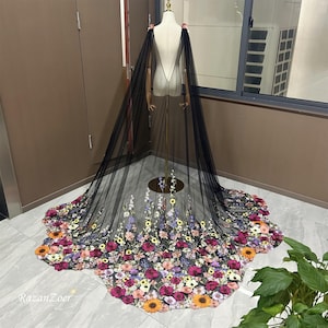 Puede incluir: Una capa de tul negro con un diseño floral. La capa está decorada con una variedad de flores coloridas, incluyendo rosa, morado, amarillo y naranja. La capa se muestra en un maniquí.