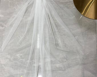 May include: A white bridal veil with a comb at the top. The veil is made of a sheer fabric and has a delicate, embellished design down the center. The veil is spread out on a light gray surface.