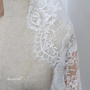 May include: Close-up of white lace fabric with floral patterns and scalloped edges. The delicate lace is draped over a mannequin, showcasing the intricate design and texture. The text "RazanZoer" is visible in the bottom left corner.