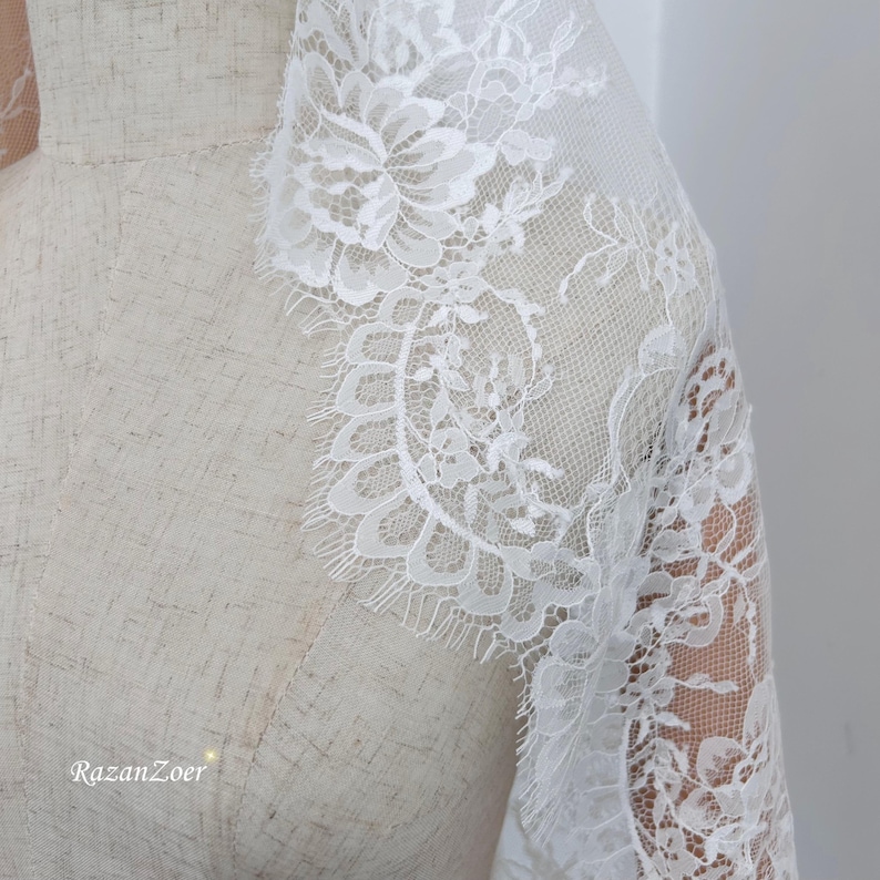 May include: Close-up of white lace fabric with floral patterns and scalloped edges. The delicate lace is draped over a mannequin, showcasing the intricate design and texture. The text "RazanZoer" is visible in the bottom left corner.