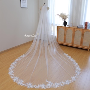 Puede incluir: Un velo de novia blanco con un delicado borde de encaje floral. El velo es largo y fluido, con una tela suave y transparente.