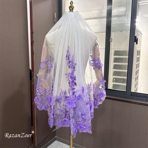 May include: A white bridal veil with intricate purple floral embroidery. The veil is displayed on a mannequin, showcasing the delicate lace and the cascading floral design. The veil is a wedding accessory.
