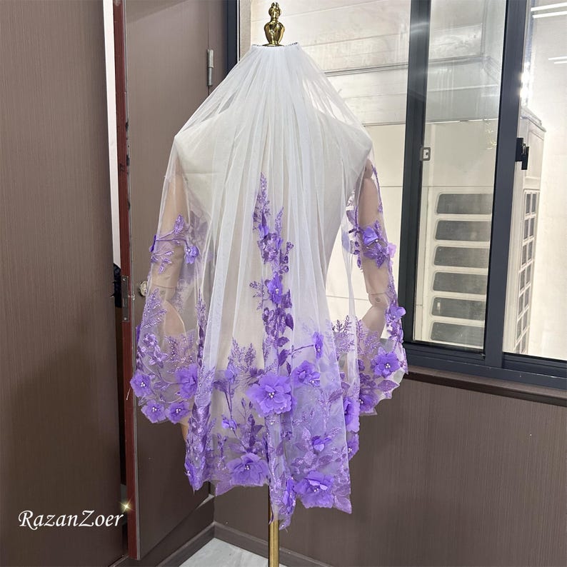 May include: A white bridal veil with intricate purple floral embroidery. The veil is displayed on a mannequin, showcasing the delicate lace and the cascading floral design. The veil is a wedding accessory.