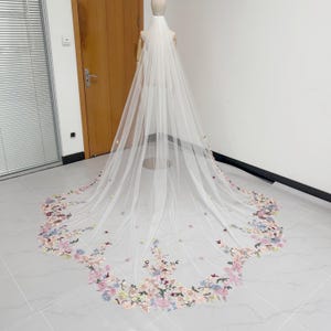 May include: A long, white bridal veil with a floral embroidered border. The veil is displayed on a mannequin and features delicate flowers in shades of pink, blue, and yellow. The veil is made of a sheer, lightweight fabric.