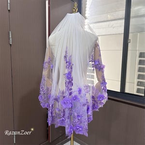May include: A bridal veil with a cascade of purple floral embellishments. The veil is sheer white, with the flowers concentrated along the lower edge and sleeves. The flowers are a vibrant shade of purple, adding a pop of color to the delicate fabric.
