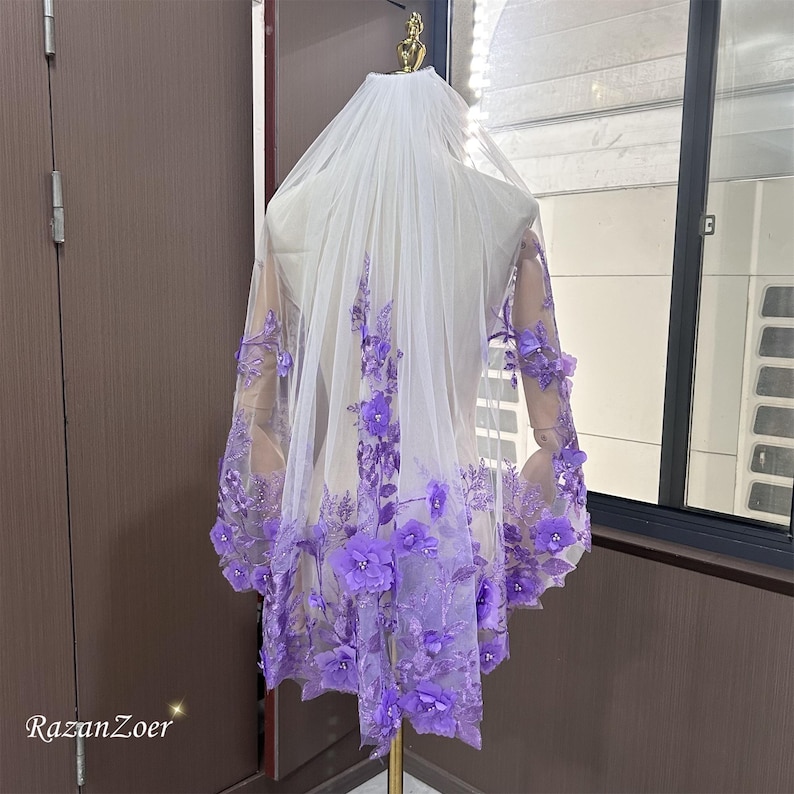 May include: A bridal veil with a cascade of purple floral embellishments. The veil is sheer white, with the flowers concentrated along the lower edge and sleeves. The flowers are a vibrant shade of purple, adding a pop of color to the delicate fabric.