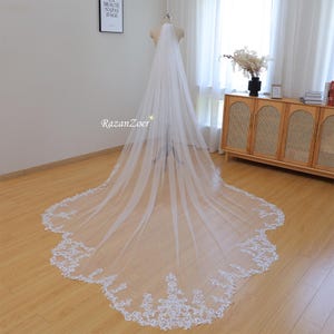 Puede incluir: Un velo de novia blanco largo con un delicado borde de encaje. El velo está hecho de tul suave y tiene un borde festoneado. El borde de encaje presenta un diseño floral.