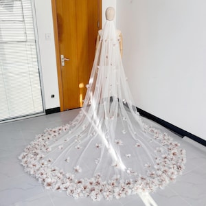 May include: A long, white bridal veil adorned with floral details. The veil is draped on a mannequin, highlighting its flowing form. The flowers are concentrated at the hem, creating a romantic aesthetic, perfect for a wedding.