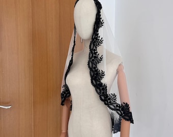 May include: A sheer white veil with a black floral lace trim. The veil is draped over a mannequin, showcasing the delicate design and texture. The black lace trim features a detailed leaf pattern, adding a touch of elegance. The brand name "RazanZoer" is visible in the bottom left corner.