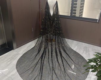 May include: A black tulle cape with gold star and crescent moon accents, draped over a mannequin. The cape features a long train and is designed for formal wear. The fabric is sheer, allowing the details to be visible.