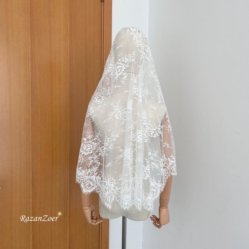 May include: A white lace veil with floral patterns. The veil is draped over a mannequin, showcasing its delicate design and texture. The veil is a bridal accessory.