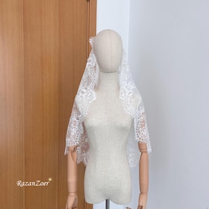 May include: A white lace veil with floral patterns is draped over a mannequin. The veil is made of delicate, sheer fabric and features intricate lace detailing along the edges. The mannequin is positioned in front of a wooden door.
