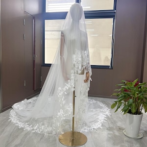 May include: A white wedding veil with floral lace detailing. The veil is displayed on a mannequin, showcasing its length and design. The veil's train extends across the floor, and the lace is visible on the edges and sleeves.
