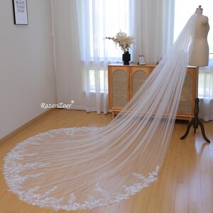 May include: A long, white bridal veil with delicate lace trim. The veil is draped over a mannequin and is displayed on a wooden floor.