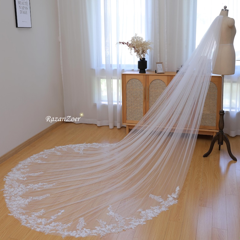May include: A long, white bridal veil with delicate lace trim. The veil is draped over a mannequin and is displayed on a wooden floor.