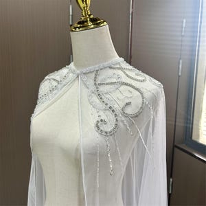 以下が含まれることがあります： 白のブライダルケープ。繊細なビーズとスパンコールの装飾が施されています。ケープは透明感のある生地でできており、キラキラ光るシルバーのアクセントが特徴です。肩と首回りのデザインが施されており、結婚式や特別な機会に最適です。