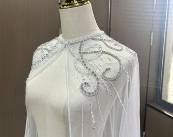 Puede incluir: Una capa de novia blanca con intrincados adornos de cuentas y lentejuelas. La capa presenta una tela delicada y transparente y está adornada con brillantes detalles plateados. El diseño incluye un detallado diseño de hombros y escote, perfecto para bodas u ocasiones especiales.