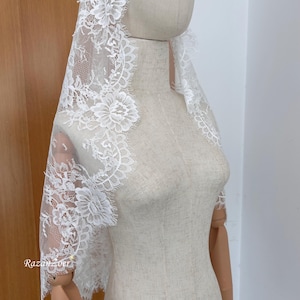 May include: A white lace veil with floral and scalloped edging is draped over a mannequin. The delicate lace features intricate floral patterns and a scalloped edge, creating an elegant and romantic look. The veil is designed to be worn over the head and shoulders.