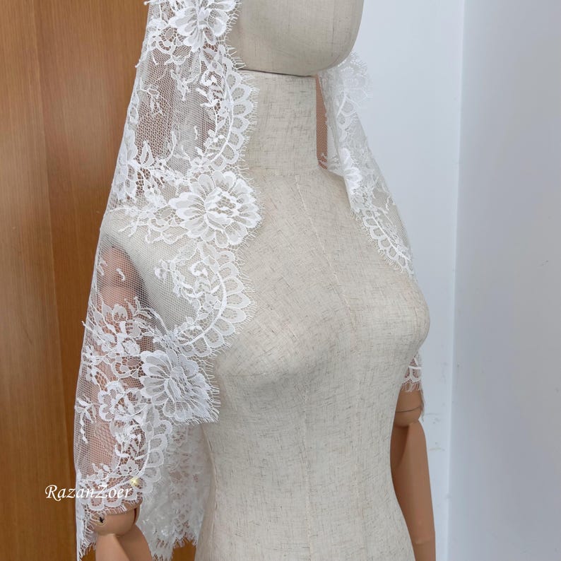 May include: A white lace veil with floral and scalloped edging is draped over a mannequin. The delicate lace features intricate floral patterns and a scalloped edge, creating an elegant and romantic look. The veil is designed to be worn over the head and shoulders.