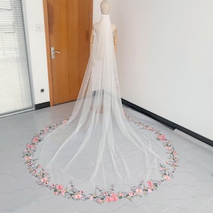 May include: A long, white bridal veil with a floral embroidered border. The veil is displayed on a mannequin, showcasing the delicate design. The embroidery features pink, blue, and green flowers.