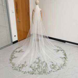May include: A long, white bridal veil with a floral lace trim in shades of green and silver. The veil is displayed on a mannequin, showcasing its length and delicate design. The veil's trim is approximately 25 cm wide.