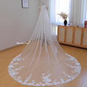 May include: A long, white tulle wedding veil with delicate floral lace trim. The veil is gathered at the top and falls in soft layers.