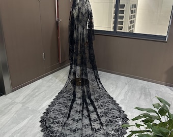May include: A long, black lace veil with a floral pattern, displayed on a mannequin. The veil features a scalloped edge and a long train. The veil is made of delicate, sheer fabric.