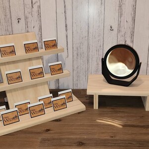 Puede incluir: Un expositor de madera con tres estantes, cada uno con peque&ntilde;as tarjetas blancas con el texto "Hey Woodcraft" impreso en negro. Un peque&ntilde;o expositor de madera con un espejo redondo negro se encuentra a la derecha del expositor.