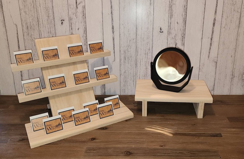 Puede incluir: Un expositor de madera con tres estantes, cada uno con peque&ntilde;as tarjetas blancas con el texto "Hey Woodcraft" impreso en negro. Un peque&ntilde;o expositor de madera con un espejo redondo negro se encuentra a la derecha del expositor.