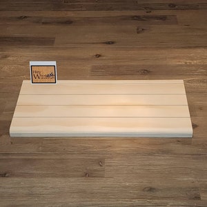 May include: A light wood shelf with a slightly rounded edge. The shelf is made of natural wood and has a light, natural finish. The shelf is sitting on a wood floor.