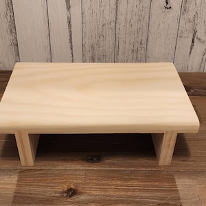 May include: A simple, natural wood riser with a rectangular top and two open sides. The riser is made of unfinished wood and has a light brown colour.