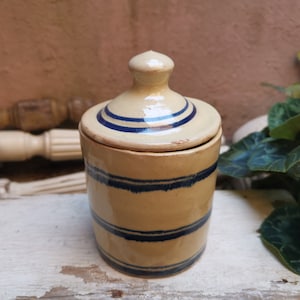 May include: A tan ceramic jar with a lid, decorated with dark blue horizontal stripes. The jar has a rounded body and a matching lid with a small knob. The design is simple and rustic, suggesting a vintage or farmhouse aesthetic.