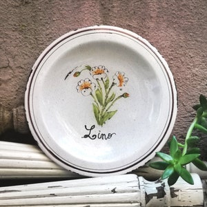 Op de afbeelding: Een wit keramiek bord met een bruine rand en een bloemenontwerp. Het bord heeft een groene steel met witte bloemen en oranje centra. Het woord "Lino" staat in het zwart op het bord.