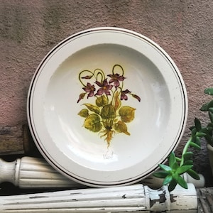 Op de afbeelding: Witte keramische plaat met een gouden rand en een handgeschilderd ontwerp van paarse viooltjes met groene bladeren. De plaat heeft het woord "Violets" in cursief op de onderkant.