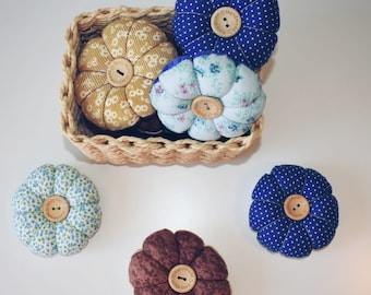 May include: Five handmade pincushions in the shape of flowers. The pincushions are made of fabric with different patterns and colors, including blue, yellow, brown, and white. Each pincushion has a wooden button in the center with the text "Handmade with love" on it.
