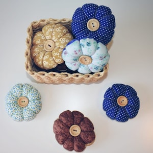 Może przedstawiać: Pięć ręcznie robionych poduszek na szpilki w kształcie kwiatów. Poduszki są wykonane z tkaniny o różnych wzorach i kolorach, w tym niebieskim, żółtym, brązowym i białym. Każda poduszka ma drewniany guzik w środku z napisem "Handmade with love".