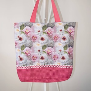 Op de afbeelding: Een roze en witte bloemen tote bag met een roze stippenbodem. De tas heeft een witte gehaakte rand en roze handvatten.