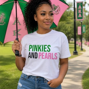 May include: A woman wearing a white t-shirt with the text "PINKIES AND PEARLS" in green and pink. She is holding a pink and green umbrella with the letters "AKA" on it. She is wearing blue jeans and is standing outside on a sunny day.
