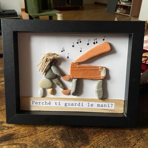 K&ouml;nnte beinhalten: Gerahmtes Kunstwerk mit einer Klavierszene aus Kieselsteinen. Eine Figur mit Seilhaar spielt Klavier, dar&uuml;ber Noten. Das Klavier besteht aus orangefarbenen und grauen Steinen. Der Text "Perch&eacute; ti guardi le mani?" steht darunter.