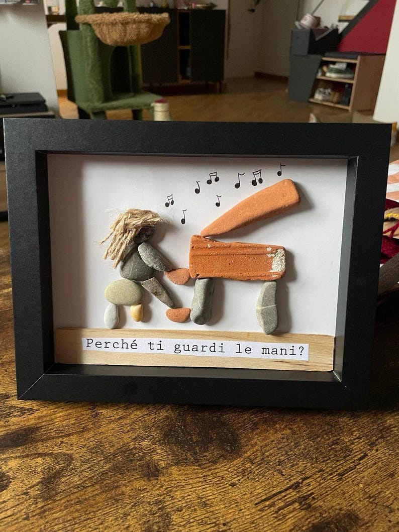 K&ouml;nnte beinhalten: Gerahmtes Kunstwerk mit einer Klavierszene aus Kieselsteinen. Eine Figur mit Seilhaar spielt Klavier, dar&uuml;ber Noten. Das Klavier besteht aus orangefarbenen und grauen Steinen. Der Text "Perch&eacute; ti guardi le mani?" steht darunter.
