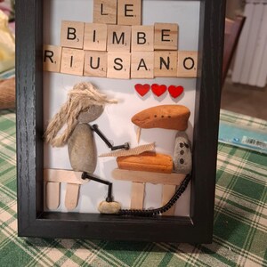 K&ouml;nnte beinhalten: Gerahmtes Kunstwerk mit den Worten "LE BIMBE RIUSANO" in Holzbuchstaben. Das Kunstwerk zeigt eine Figur aus Steinen und Seilen, mit drei roten Herzakzenten und anderen Stein- und Holzelementen.