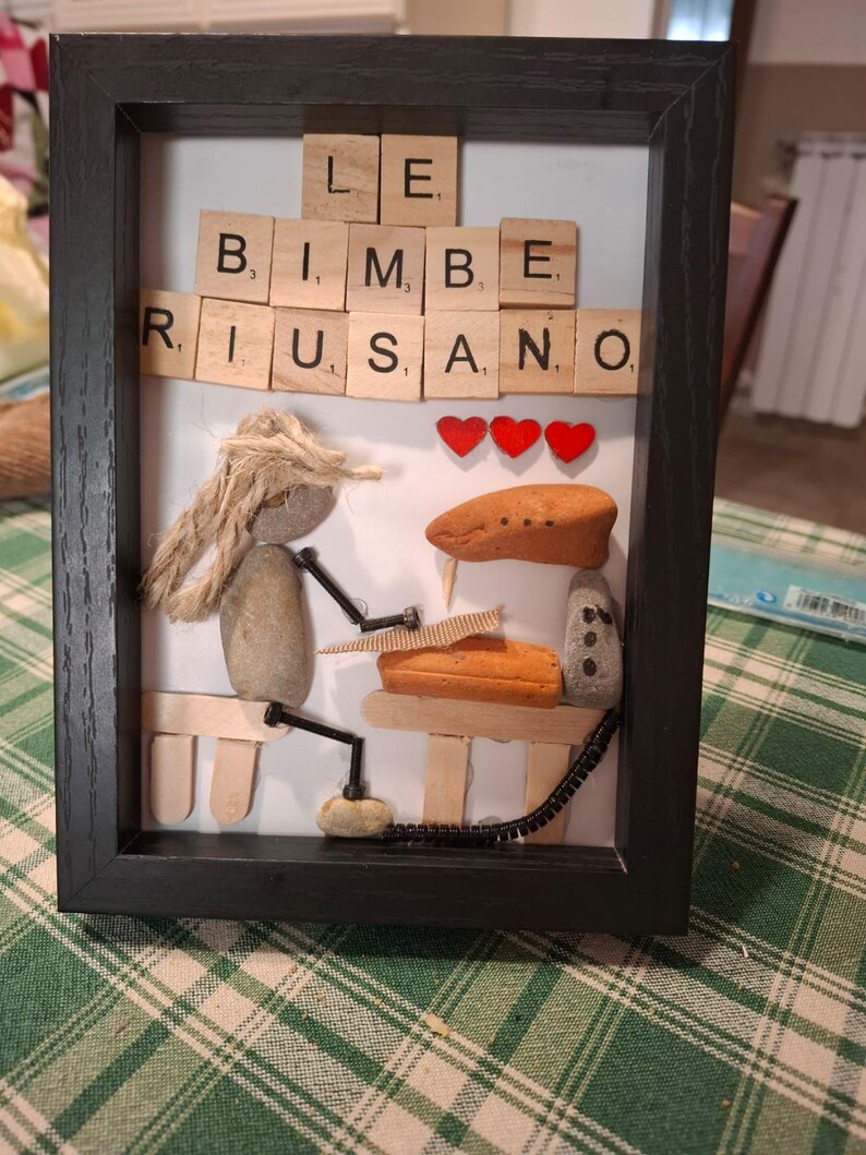 K&ouml;nnte beinhalten: Gerahmtes Kunstwerk mit den Worten "LE BIMBE RIUSANO" in Holzbuchstaben. Das Kunstwerk zeigt eine Figur aus Steinen und Seilen, mit drei roten Herzakzenten und anderen Stein- und Holzelementen.