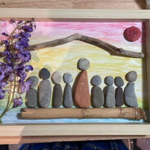 K&ouml;nnte beinhalten: Gerahmtes Kunstwerk mit einer Familie aus Steinen. Der Hintergrund ist mit einem Sonnenuntergang und einer Sonne bemalt. Lila Blumen und ein St&uuml;ck Treibholz sind ebenfalls enthalten. Der Rahmen besteht aus hellem Holz.