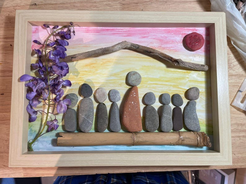 K&ouml;nnte beinhalten: Gerahmtes Kunstwerk mit einer Familie aus Steinen. Der Hintergrund ist mit einem Sonnenuntergang und einer Sonne bemalt. Lila Blumen und ein St&uuml;ck Treibholz sind ebenfalls enthalten. Der Rahmen besteht aus hellem Holz.
