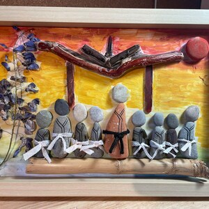Può includere: Opera d'arte incorniciata con una scena di figure fatte di pietre, una struttura in legno e uno sfondo colorato. Le figure sono disposte in fila, alcune con nastri bianchi. L'opera è incorniciata in una cornice di legno chiaro.