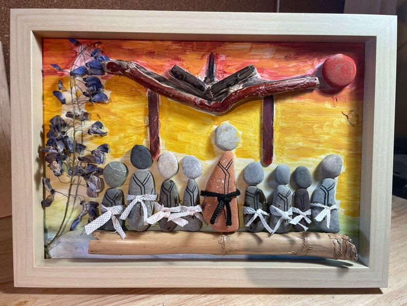 K&ouml;nnte beinhalten: Gerahmtes Kunstwerk mit einer Szene aus Steinfiguren, einer Holzstruktur und einem farbenfrohen Hintergrund. Die Figuren sind in einer Reihe angeordnet, einige tragen wei&szlig;e B&auml;nder. Das Kunstwerk ist in einem hellen Holzrahmen gefasst.