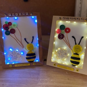 Può includere: Due opere d'arte incorniciate a tema ape. Ognuna presenta un'ape gialla e nera con palloncini a bottone e luci fatate. La parola "AURORA" è in cima a ogni cornice. La data "20 SETTEMBRE 2023" è in basso.