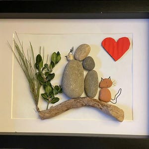 K&ouml;nnte beinhalten: Gerahmtes Kunstwerk mit einer Familienszene aus nat&uuml;rlichen Materialien. Glatte Steine bilden Figuren, ein rotes Papierherz und eine Katze. Gr&uuml;nes Laub und Treibholz verleihen der Komposition Textur.
